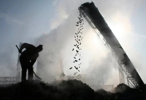 Worker shovels coal beneath a coal conveyer gantry, in front of hazy smoke-filled background.
