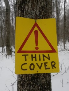 Yellow plastic sign on tree with snow in the background. Sign is marked 'Thin Cover' beneath triangular road marking symbol for warning.