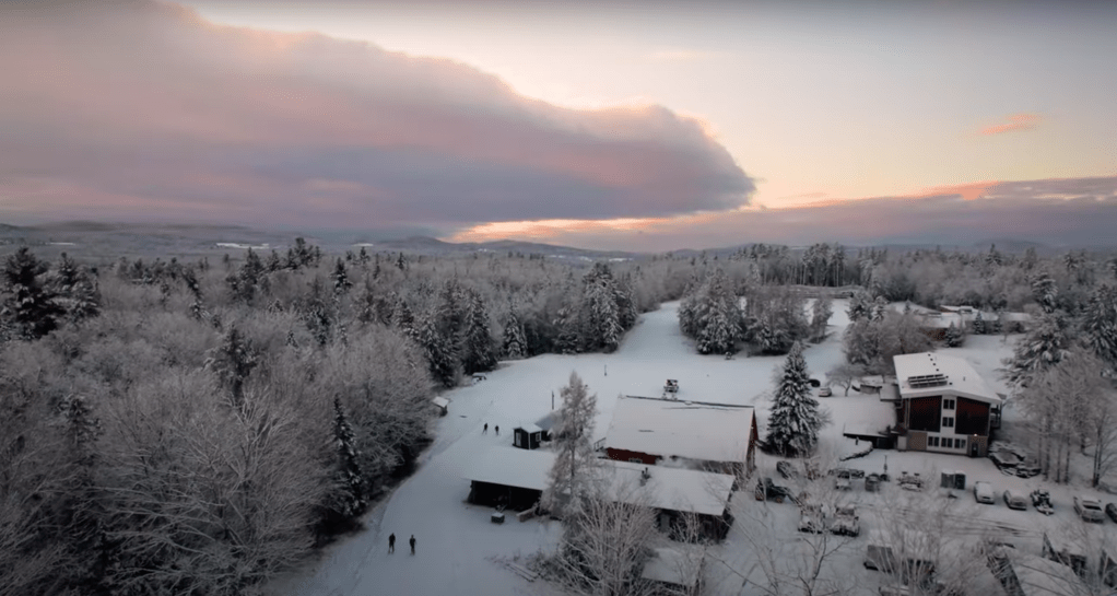 Screenshot from aerial footage showing Craftsbury Outdoor Center campus with snow on trees and on the ground. From '90 DAYS TIL SNOW - A Fall with the University of New Hampshire Ski Team' 