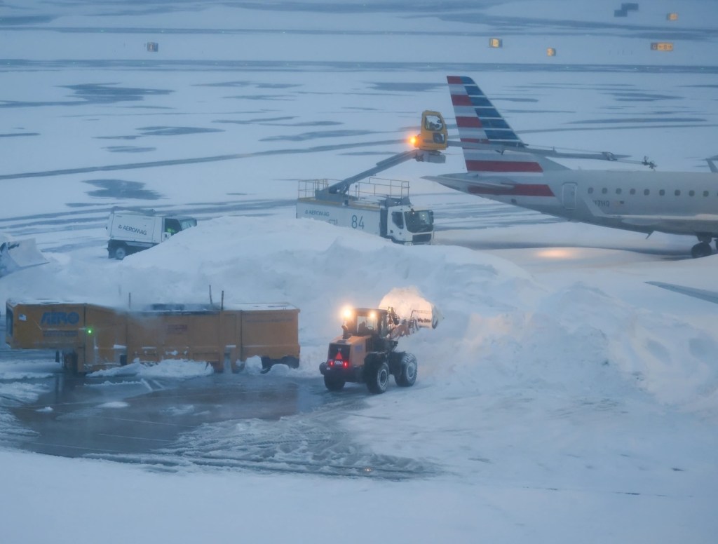 Bulldozers dump snow into an ice melter near an American Airlines plane at LaGuardia Airport (LGA) during a winter storm in the Queens borough of New York, US, on Sunday, Jan. 25, 2026. A massive winter storm reached the US Atlantic Coast on Sunday, bringing heavy snow and ice, straining electric grids and grounding thousands of flights at levels not seen since the pandemic. Photographer: Michael Nagle/Bloomberg